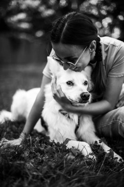 Beyaz tüylü köpeğini çimenlikteki parkta kucaklayan gülümseyen kadının siyah beyaz fotoğrafı. Evlat edinme, kurtarılma, barınma, yoldaş, hayvan, en iyi arkadaş.