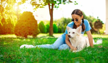 Parkta çimlerin üzerinde sevimli beyaz tüylü köpeğini kucaklayan mutlu, genç, güzel esmer kız. Evlat edinme, kurtarılma, barınma, yoldaş, evcil hayvan.