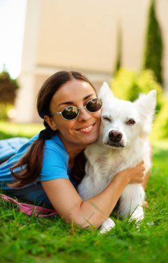 Parkta çimlerin üzerinde sevimli beyaz tüylü köpeğini kucaklayan mutlu, genç, güzel esmer kız. Evlat edinme, kurtarılma, barınma, yoldaş, evcil hayvan.