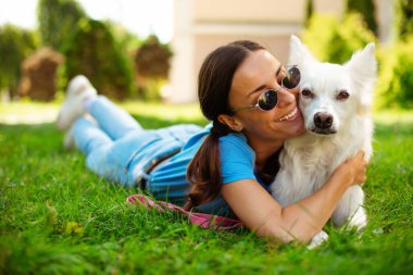 Parkta çimlerin üzerinde sevimli beyaz tüylü köpeğini kucaklayan mutlu, genç, güzel esmer kız. Evlat edinme, kurtarılma, barınma, yoldaş, evcil hayvan.