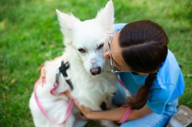 Parkta çimlerin üzerinde sevimli beyaz tüylü köpeğini kucaklayan mutlu, genç, güzel esmer kız. Evlat edinme, kurtarılma, barınma, yoldaş, evcil hayvan.