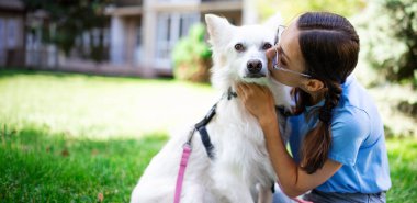 Mutlu kadın çimenlikteki parkta beyaz tüylü köpeğine sarılıp öpüyor. Evlat edinme, kurtarılma, barınma, yoldaş, hayvan, en iyi arkadaş.