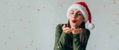 Noel şapkalı heyecanlı genç bir kadının yakın plan fotoğrafı ellerinden konfeti üflüyor ve yeni yılı partiyle kutluyor.