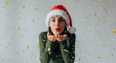 Noel şapkalı heyecanlı genç bir kadının yakın plan fotoğrafı ellerinden konfeti üflüyor ve yeni yılı partiyle kutluyor.