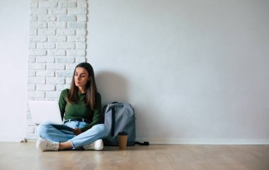 Gündelik giysiler içinde genç, kendine güvenen bir bayan bir dizüstü bilgisayar kullanıyor ve gri arka planda yerde otururken gülümsüyor. Eğitim, eğitim, üniversite, üniversite, mezuniyet konsepti