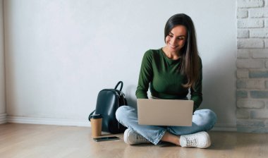 Gündelik giysiler içinde genç, kendine güvenen bir bayan bir dizüstü bilgisayar kullanıyor ve gri arka planda yerde otururken gülümsüyor. Eğitim, eğitim, üniversite, üniversite, mezuniyet konsepti