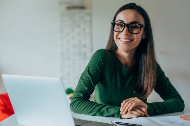 Genç, mutlu, pozitif, gözlüklü, serbest çalışan güzel iş kadınının portresi. Ev ofisinin içinde dizüstü bilgisayar kullanarak otur.