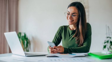 Evde dizüstü bilgisayarla çalışırken akıllı telefon kullanan kendine güvenen genç iş kadınının portresini kapat. 