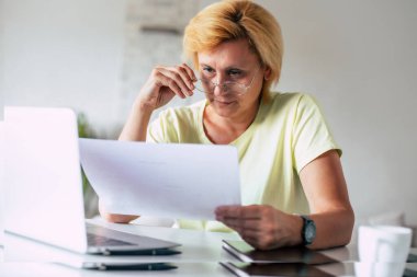 Bilgisayarlı ve belgeli orta yaşlı bir kadının yakın plan çekimi, gözlüklü bir kadın ev idaresinde kağıt fatura ödemesi yapıyor banka finansmanı, kredi emekliliği ödemesi planlayan vergileri hesaplıyor.