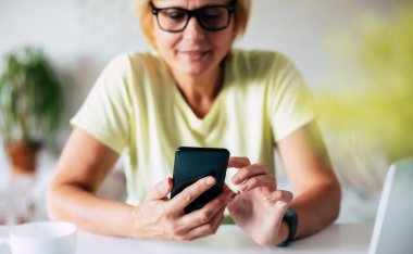  Cep telefonuyla sörf sitesi. İnternetten alışveriş yaparken elinde akıllı telefonla fotoğraf çeken orta yaşlı bir kadın. Gözlüklü, gülümseyen bir kadın. Mesajlaşıyor ya da telefonda sohbet ediyor.