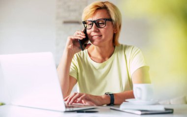 Ofiste çalışırken elinde akıllı telefonla konuşan ve internette sörf yapan kendinden emin olgun bir kadın. Dizüstü bilgisayar kullanan gözlüklü bir kadın.