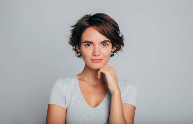 Gülümseyen, kısa saçlı, sıradan giyinmiş, sakince kameraya bakan mutlu bir kadının portresini yakından çek. Boş gri duvarda izole edilmiş göz kamaştırıcı kızın stüdyo fotoğrafı..