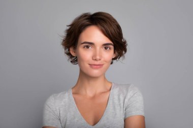 Gülümseyen, kısa saçlı, sıradan giyinmiş, sakince kameraya bakan mutlu bir kadının portresini yakından çek. Boş gri duvarda izole edilmiş göz kamaştırıcı kızın stüdyo fotoğrafı..