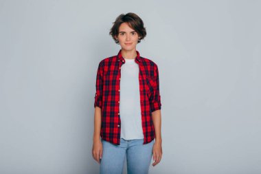 Kısa kıvırcık saçlı, gri arka planda kareli gömlekli genç, ciddi bir kadın kameraya bakıyor.