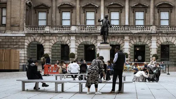 Londra, İngiltere, 30 Nisan 2023: Aydınlık bir günde insanlar dışarıda bronz bir heykelin yanında oturuyor