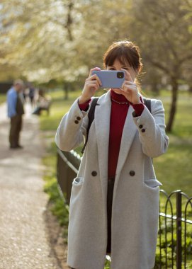 Londra, İngiltere, 25 Nisan 2023: İspanyol Kadın telefonuyla fotoğraf çekiyor, parkta, kameraya bakıyor..