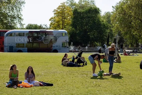 Marble Arch 'ın yanındaki çimlerin üzerinde oturan insanlar arka planda çift katlı bir Loewe markası var.