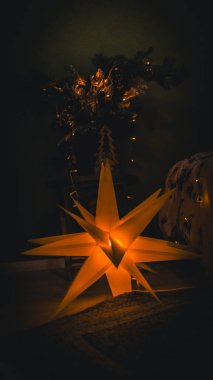 Güzel aydınlatılmış yıldız şekilli bir dekorasyon loş ışıklı bir odada sıcak bir parıltı saçarak hoş bir festival atmosferi yaratır. Arka planda, peri ışıkları parıldıyor, tatil ruhunu güçlendiriyor. Yumuşak ışıklandırma ve zengin altın renkler