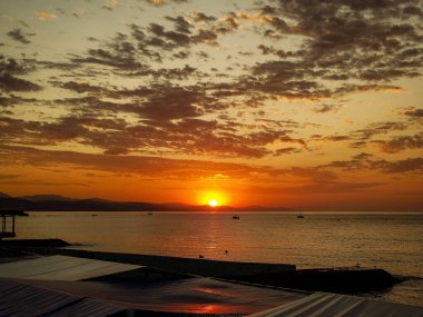 Büyüleyici bir altın gün batımı gökyüzünü ve denizi sıcak renklerle boyar. Bir balıkçı bir iskelede dikilirken bir yelkenli uzaklarda sürükleniyor, sakin ve güzel bir manzara yaratıyor..