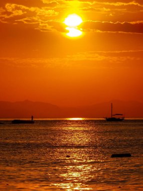 Büyüleyici bir altın gün batımı gökyüzünü ve denizi sıcak renklerle boyar. Bir balıkçı bir iskelede dikilirken bir yelkenli uzaklarda sürükleniyor, sakin ve güzel bir manzara yaratıyor..