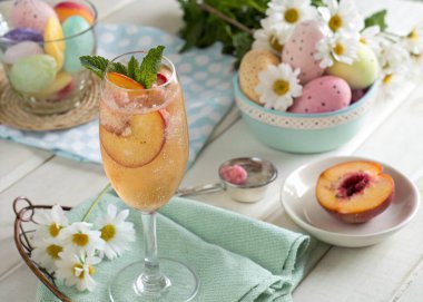 A sparkling peach Bellini in a tall champagne flute is garnished with a slice of fresh peach and a sprig of mint. The pastel Easter theme is enhanced by the surrounding decorations of colorful eggs, fresh daisies, and a soft tablecloth in spring hues clipart