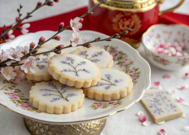 A photo of a traditional tea setting with a focus on pastries. The main objects include a white plate with pastries featuring floral designs, a red and gold teapot, and a cup with a similar decorative theme. The plate is on a table with a white base, clipart