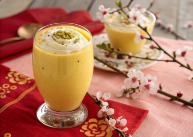 A photo of a table setting with a creamy, orange-colored drink garnished with a dollop of white cream and a touch of green herbs. The drink is served in a tall glass and is on a table adorned with a rich red cloth featuring a traditional golden patte clipart