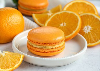 A close-up photo of a culinary scene featuring a macaron-like pastry and slices of fresh orange fruit. The macaron is golden-brown and has a layered structure on a white plate. The orange slices, with their vibrant color and segmented interior, are s clipart