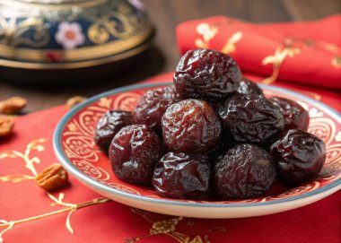 A photo featuring a close-up view of a plate of dried dates, arranged closely together on a decorative table setting. The dates have a rich, dark brown color and a glossy sheen. The table setting has a red cloth with intricate gold patterns and a bac clipart