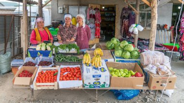 Osh, Kırgızistan - Mayıs 2022: Kırgızistan 'daki hareketli bir yerel pazarda geleneksel kıyafet giymiş Kırgız kadın satıcılar, yoldan geçenlere taze ve renkli meyve ve sebzelerini gururla sergiliyor.