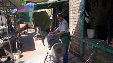 Osh, Kyrgyzstan - May 2022: Man sharpening a knife with a grinder at a local blacksmith shop with spark fireworks. Laborer using grinding equipment.