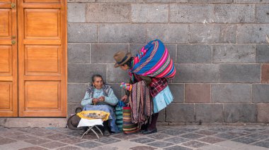 Cusco, Peru - 24 Ekim 2023: Perulu kadınlar sokakta taş bir duvarın önünde oturan turistler için hediyelik eşya ve yiyecek satıyor