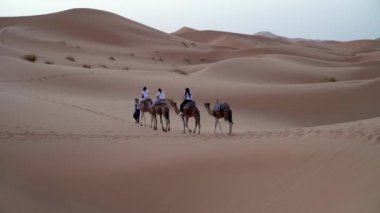 Sahra Çölü, Fas - 10 Kasım 2023: Kum tepeleri üzerinde deve sürüşü yapan turistler, klasik bir Sahra Çölü turizmi deneyimi olan Berber bir adamın rehberliğinde