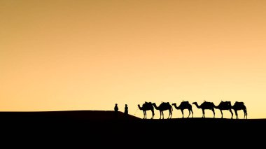 Sahra Çölü, Fas 'ta gün batımında kum tepeleri üzerinde deve kervanlarıyla kimliği belirsiz Berberilerin silueti.
