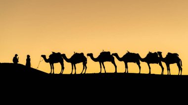 Sahra Çölü, Fas 'ta gün batımında kum tepeleri üzerinde deve kervanlarıyla kimliği belirsiz Berberilerin silueti.