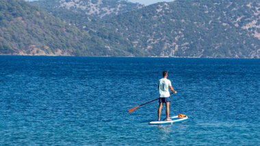 Mugla, Turkey - 11 Oct 2024: Man enjoying a stand-up paddleboarding adventure on the crystal-clear waters of Sarsala Bay, in the beautiful Mediterranean, embracing an active vacation lifestyle clipart