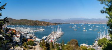 Fethiye, Türkiye - 3 Kasım 2024: Fetiye Yat Marina 'nın turkuaz Akdeniz' de demirli, yemyeşil dağlar ve canlı bir kıyı kasabasıyla çevrili yelkenli ve lüks yatları ile havadan görünüşü.