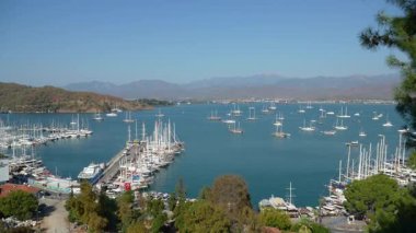 Fethiye, Türkiye - 3 Kasım 2024: Fetiye Yat Marina 'nın turkuaz Akdeniz' de demirli, yemyeşil dağlar ve canlı bir kıyı kasabasıyla çevrili yelkenli ve lüks yatları ile havadan görünüşü.