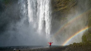 İzlanda 'daki güçlü Skogafoss şelalesinin altında, sisli ve canlı bir gökkuşağı tarafından çerçevelenmiş kimliği belirsiz bir kadın duruyor.