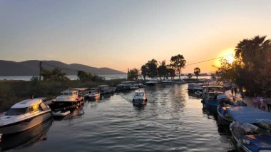 Akyaka, Türkiye - 9 Ekim 2024: Azmak nehrinin Akdeniz 'le buluştuğu Akyaka kentinin manzaralı gün batımı manzarası, kanal boyunca demirlemiş balıkçı tekneleri, yatlar, palmiye ağaçları ve altın saat ışığında dağlarla