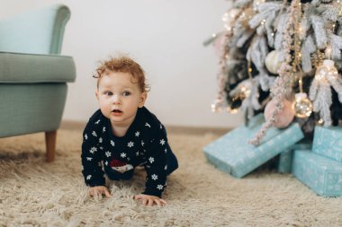 Geleneksel Noel süveteri giyen kıvırcık bir çocuk Noel ağacının yanında vakit geçiriyor.