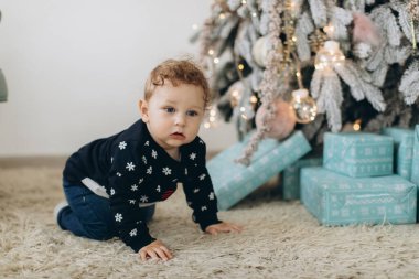 Geleneksel Noel süveteri giyen kıvırcık bir çocuk Noel ağacının yanında vakit geçiriyor.