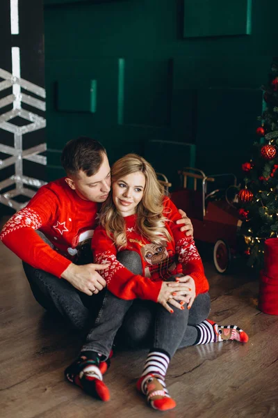 伝統的なクリスマスの赤いセーターで魅力的なカップルはクリスマスツリーの近くで時間を過ごす — ストック写真