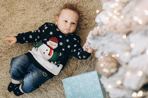 伝統的なクリスマスセーターの中の小さな巻き男の子はクリスマスツリーの近くの家で時間を過ごす — ストック写真