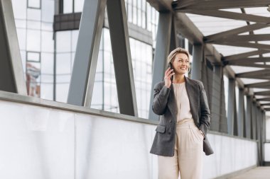 Takım elbiseli, gri ceketli, modern şehir ve ofis binalarında gülümseyen ve konuşan güzel bir iş kadınının portresi.