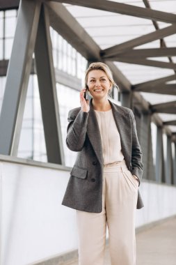 Takım elbiseli, gri ceketli, modern şehir ve ofis binalarında gülümseyen ve konuşan güzel bir iş kadınının portresi.