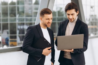 Takım elbiseli iki yakışıklı genç iş adamı bir dizüstü bilgisayar ve bir klasör tutuyorlar ve bir çalışma projesini tartışıyorlar ya da kentsel ofis binalarının arka planına karşı video görüşmesi yapıyorlar..