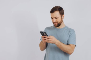 Modern çağın sakallı, akıllı bir adamın portresi. Akıllı bir telefon tutuyor ve kablosuz kulaklıkla fotokopi çekilen beyaz bir arka planda bir şeyler dinliyor.