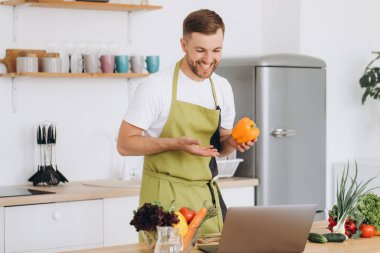 Evde mutlu bir adamın portresi, sebze salatası yapan bir adam kameraya bakıyor ve gülümsüyor, sebzeleri dilimliyor, dizüstü bilgisayarla çevrimiçi pişirme eğitimi alıyor.