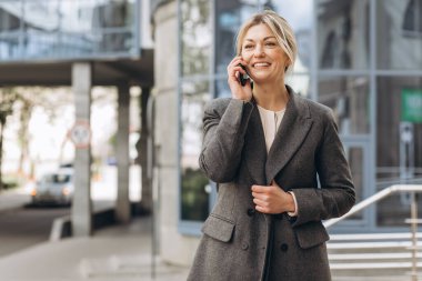 Takım elbiseli, gri ceketli, modern şehir ve ofis binalarında gülümseyen ve konuşan güzel bir iş kadınının portresi.